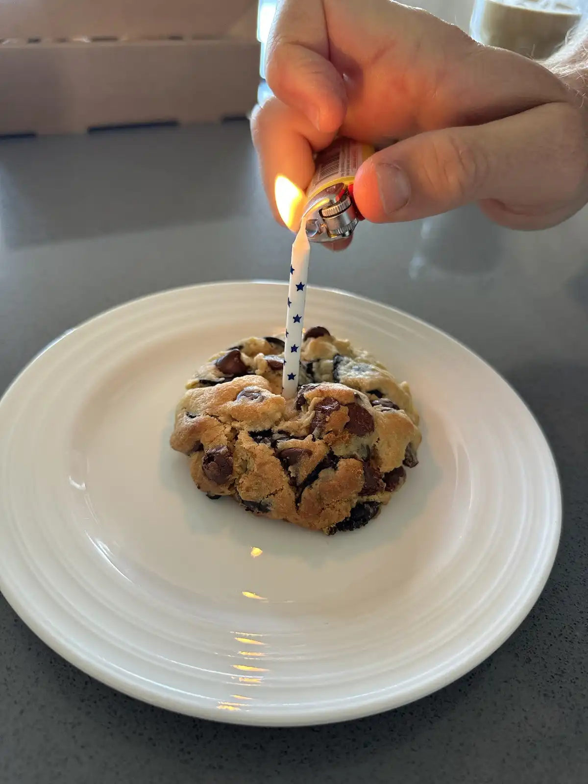 A hand holds a lighter and lights a blue star-patterned birthday candle stuck in a chocolate chip cookie on a white plate…