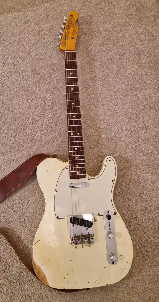 A vintage, heavily worn white Fender Telecaster electric guitar with a brown leather strap lies on a beige carpet. The…