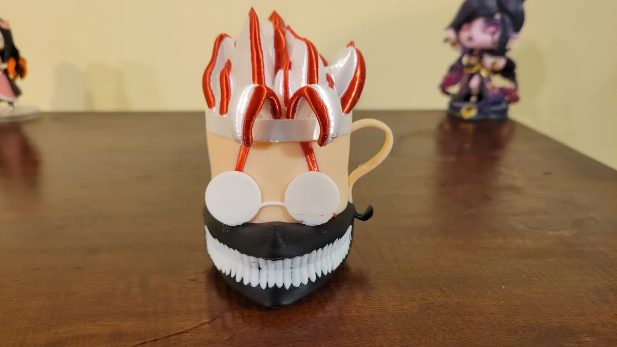 A 3D-printed mug is decorated to resemble a cartoonish, menacing face with spiky red and white hair, round white glasses…