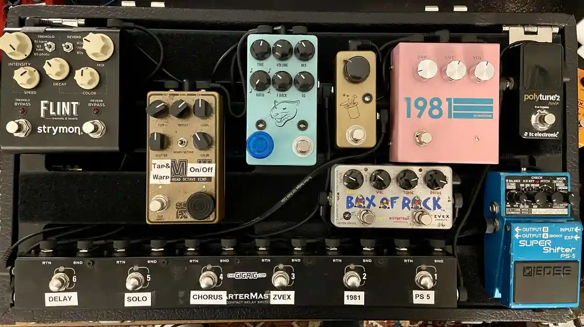 A close-up photo of a guitar pedalboard filled with various effects pedals, including a pink '1981' pedal, a blue 'Box of…
