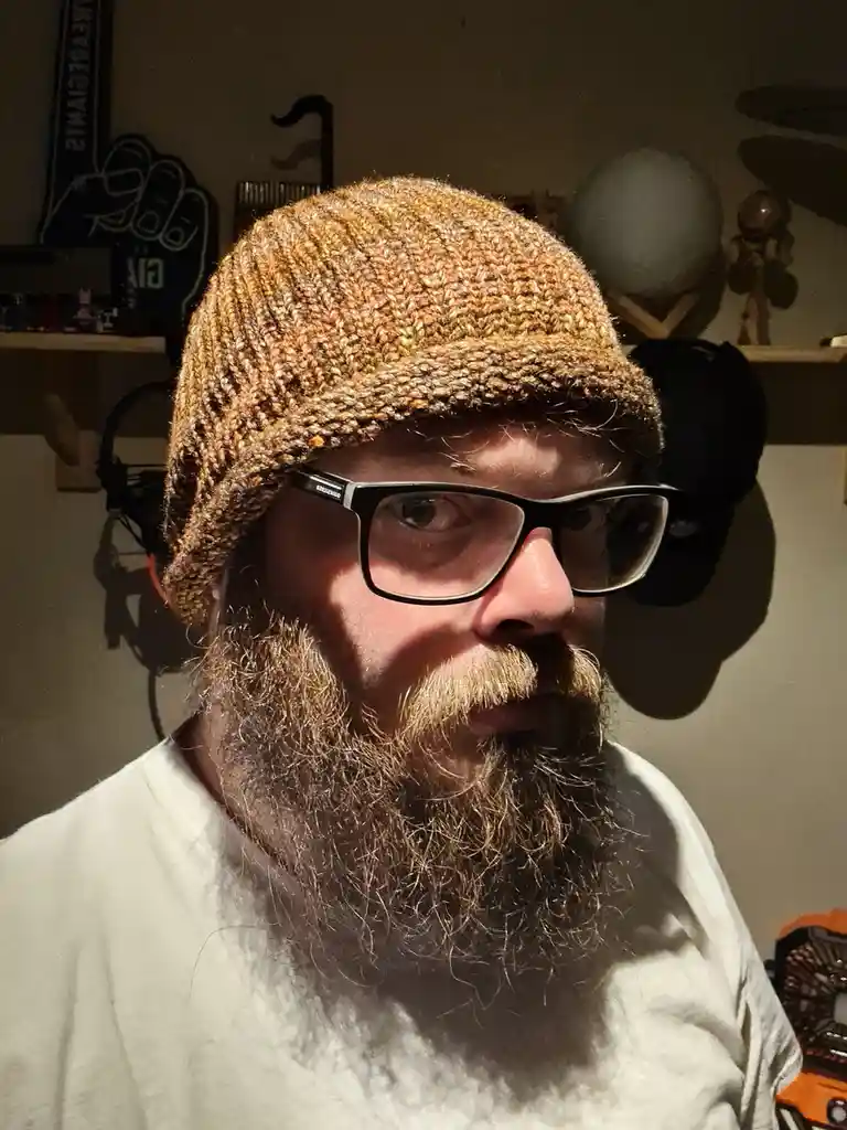 A man with a full beard, glasses, and a brown knit beanie stares intently at the camera. Behind him, shelves hold various…