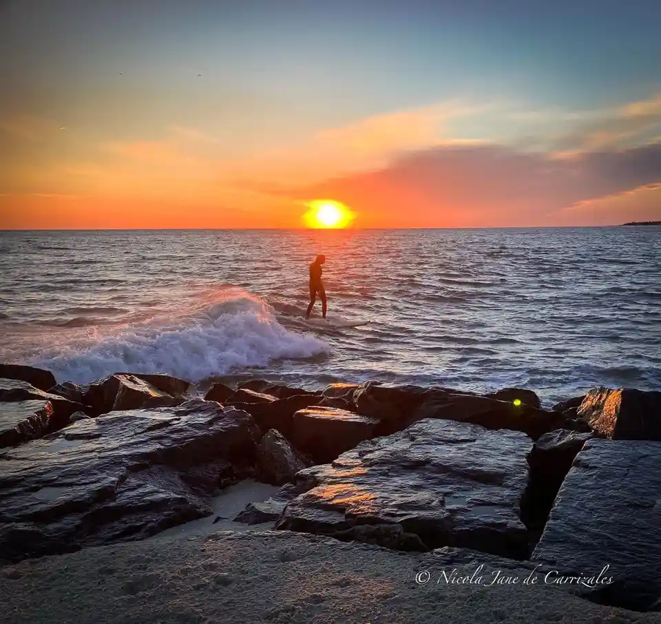 A person is silhouetted on a surfboard in the ocean during a vibrant sunset, with waves breaking near dark rocks in the…