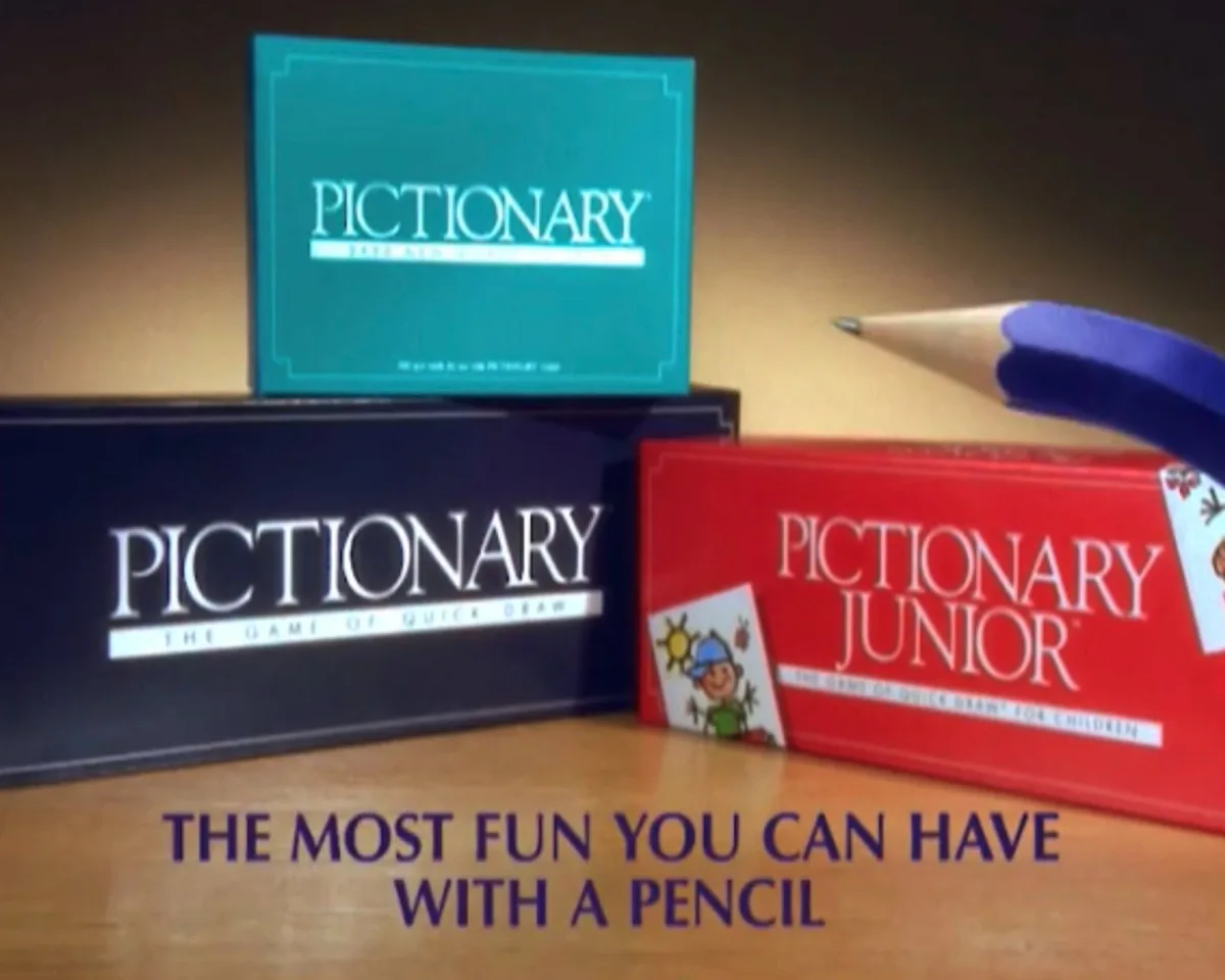 Three Pictionary game boxes — teal, black, and red — are arranged on a wooden surface. A large, stylized pencil with a…