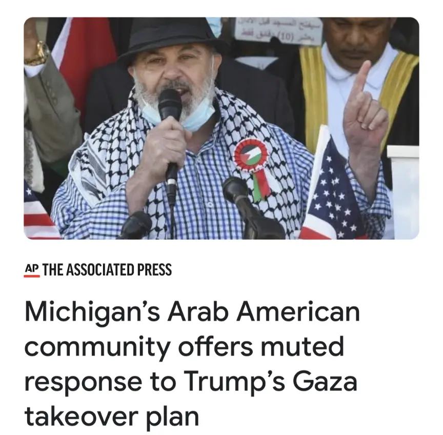 A man wearing a black hat, a checkered keffiyeh, and a face mask speaks into a microphone while holding an American flag. He…