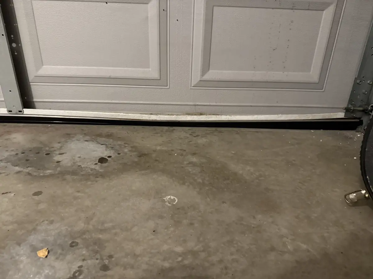 A low-angle photo shows the bottom of a white garage door with a black rubber seal against a stained concrete floor. The…