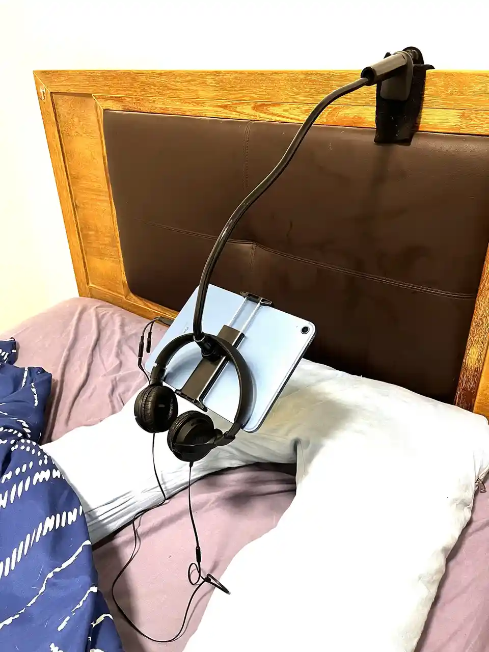 A light blue tablet is mounted on a flexible black arm attached to a wooden headboard with a brown leather panel. The tablet…