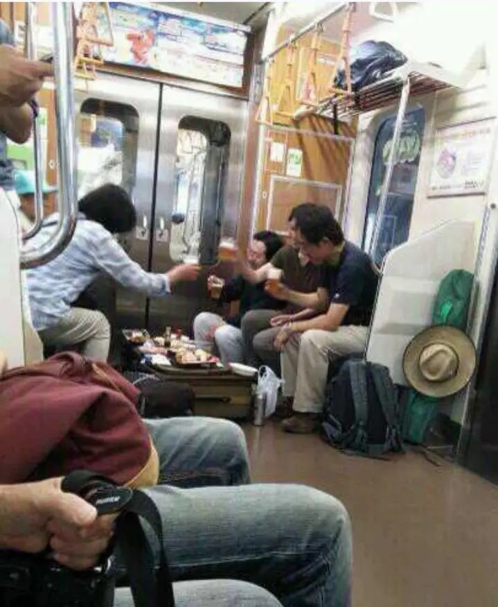 Inside a crowded subway car, a group of people are seated on the floor around an open suitcase filled with food and drinks…