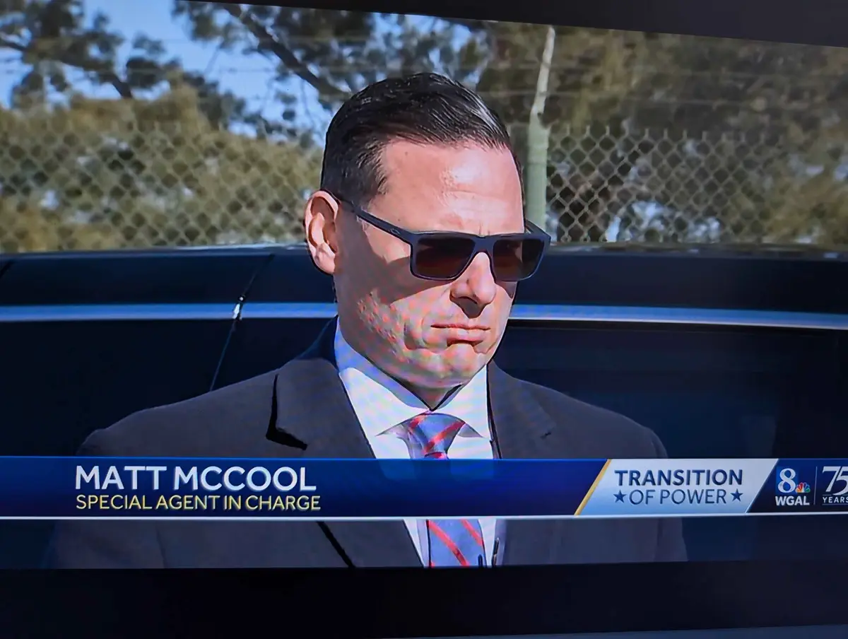 A man in a suit and sunglasses stands outdoors in front of a dark vehicle with a chain-link fence behind him. A news graphic…