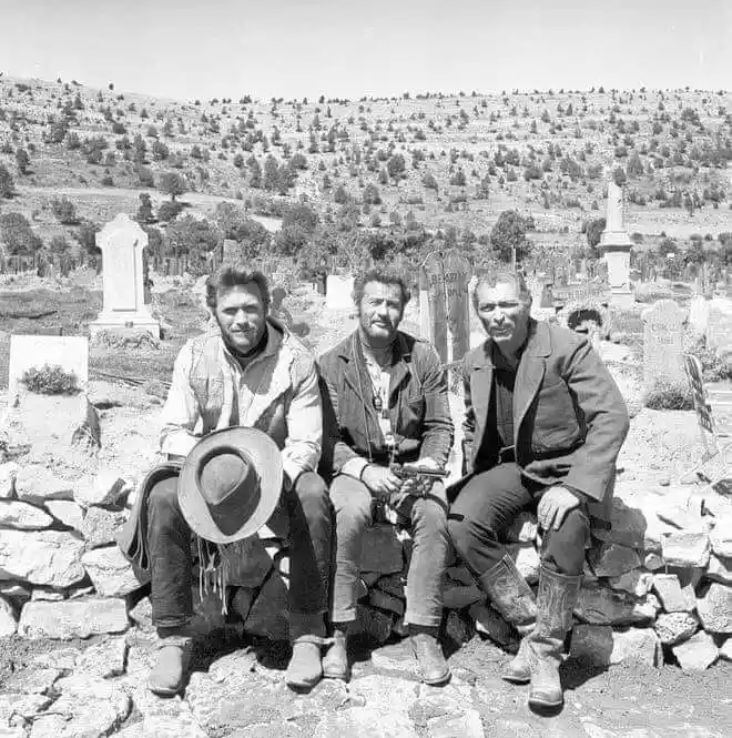 Three men in cowboy attire sit on a low stone wall in a cemetery, surrounded by gravestones under a bright sky. The man on…
