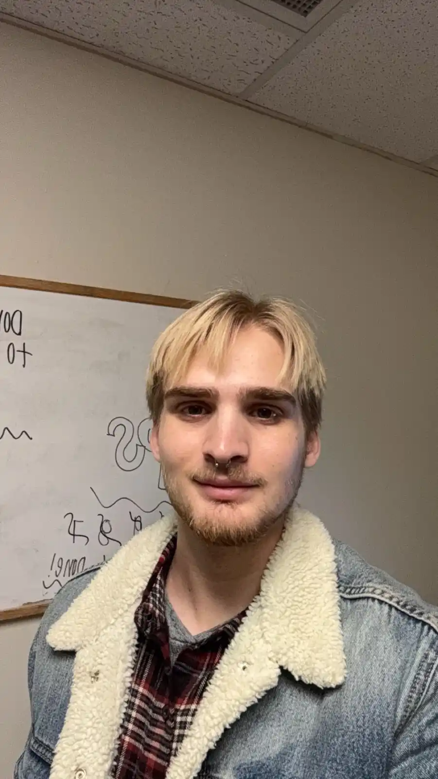 A young man with blonde hair, a nose ring, and a goatee smiles at the camera while wearing a denim jacket with a white…