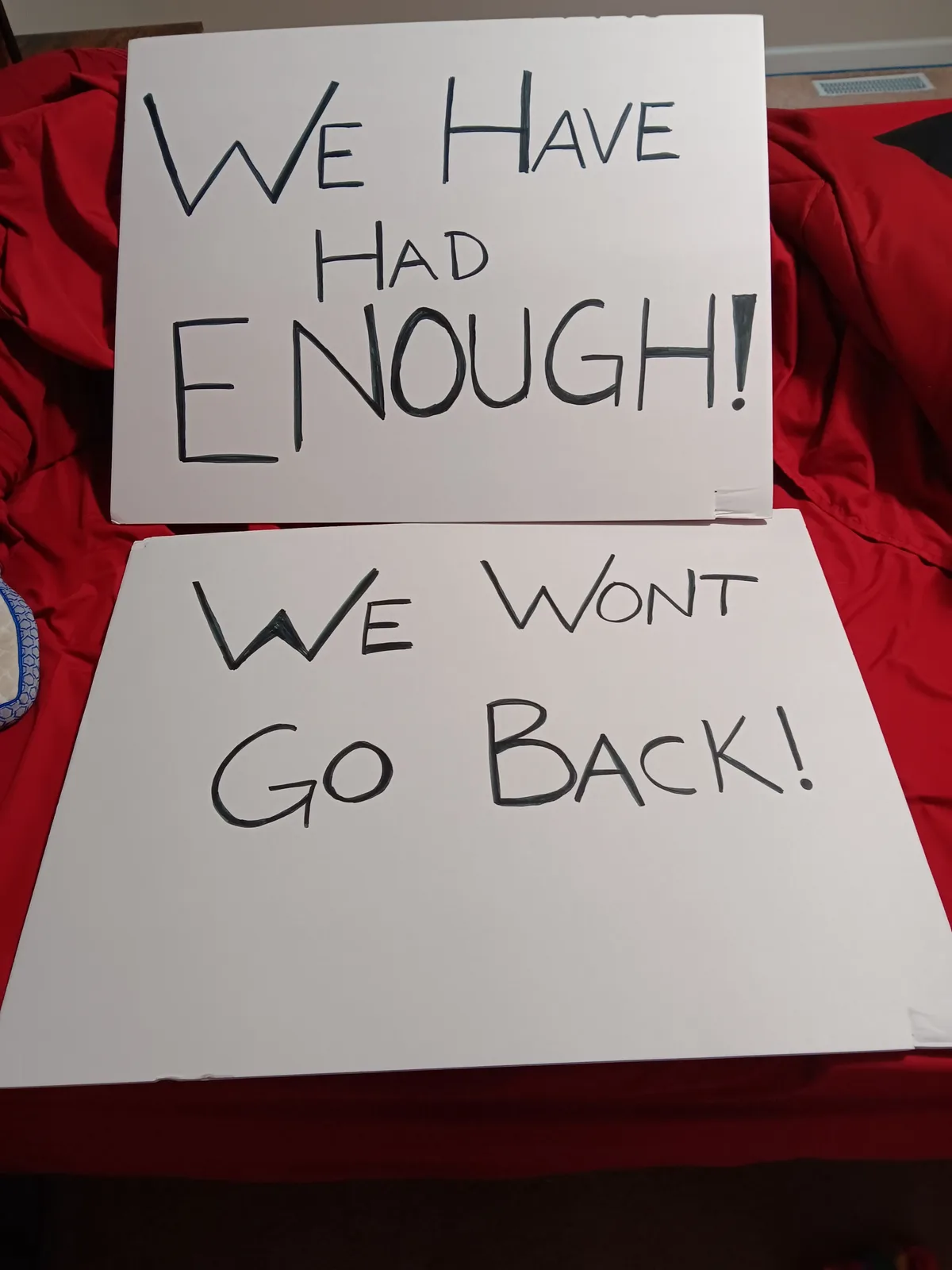 Two handmade protest signs with bold, black marker text are placed on a red bedspread. The top sign reads 'WE HAVE HAD…