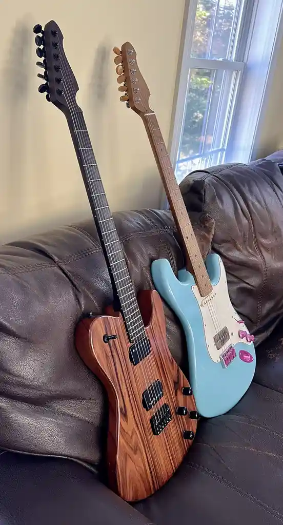 Two electric guitars lean against each other on a brown leather couch near a window. One is a dark wood with a natural grain…