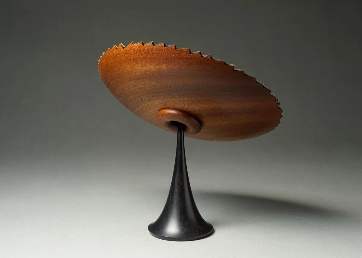 A wooden, saucer-shaped object with a serrated edge sits atop a black, conical stand against a gray background. The overlaid…
