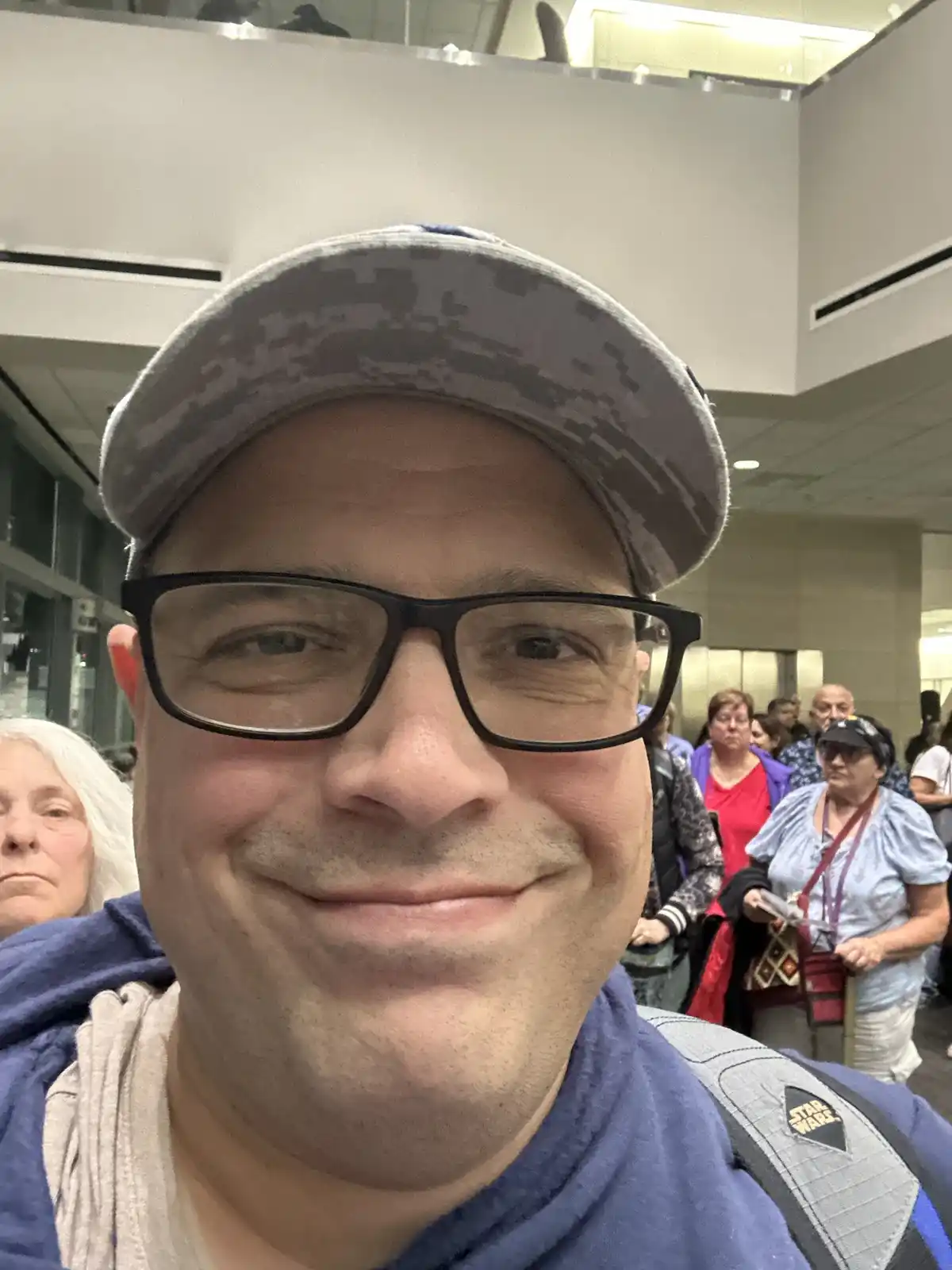 A man wearing a camouflage baseball cap and black-rimmed glasses smiles at the camera while taking a selfie in a crowded…