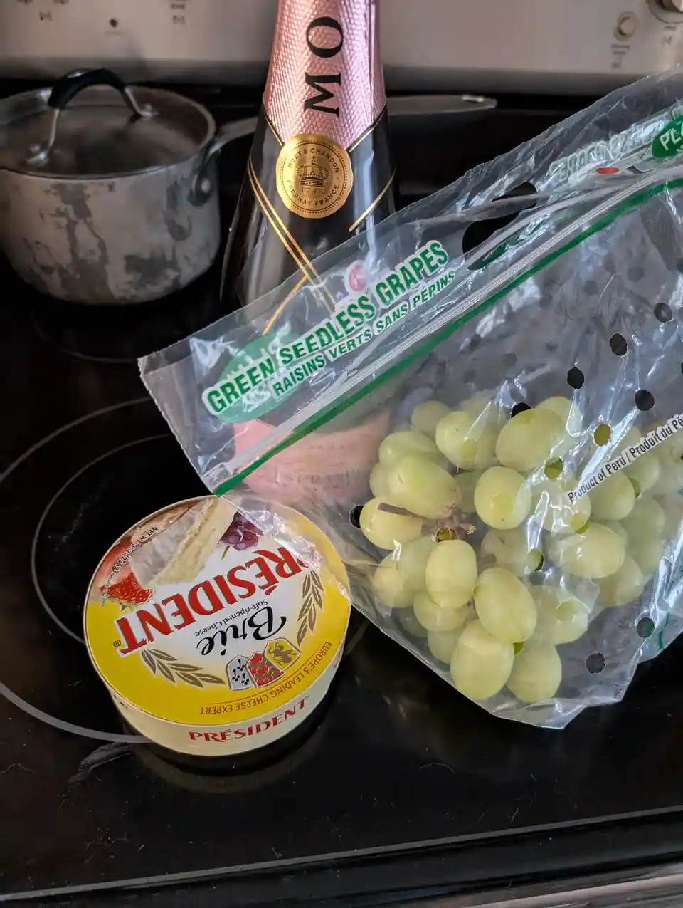 A close-up photo shows a bottle of Moët & Chandon champagne, a bag of green seedless grapes, and a round container of…