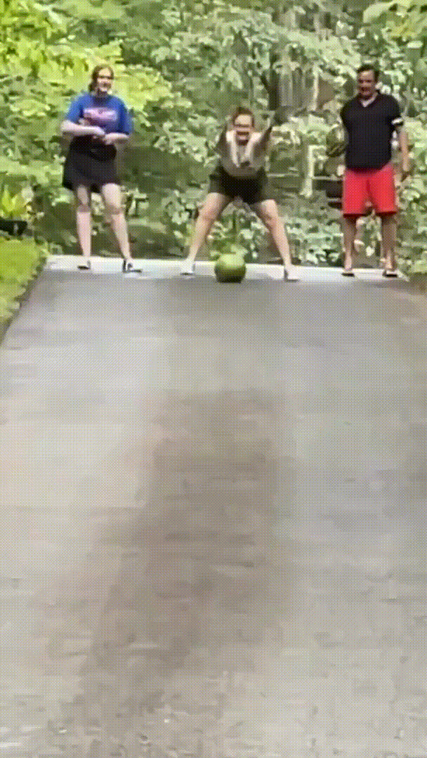 Three people stand at the top of a paved hill, watching a watermelon roll toward them. The watermelon rolls down the hill…