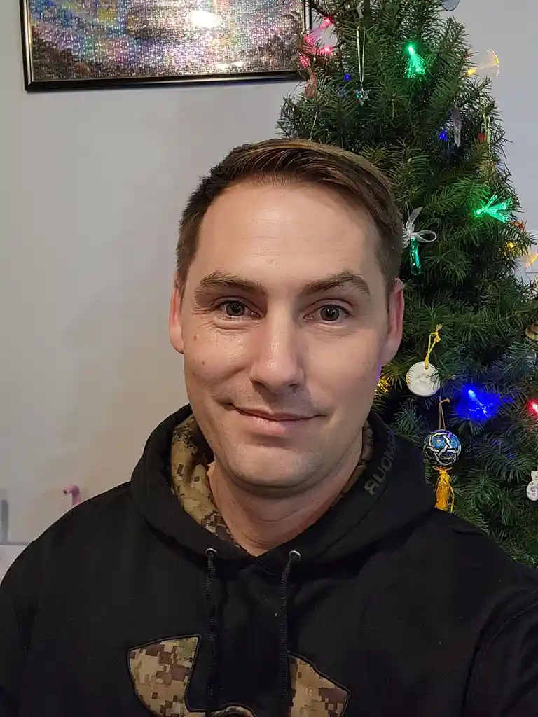 A man with short brown hair and a slight smile wears a black hoodie with a digital camouflage pattern on the collar and…