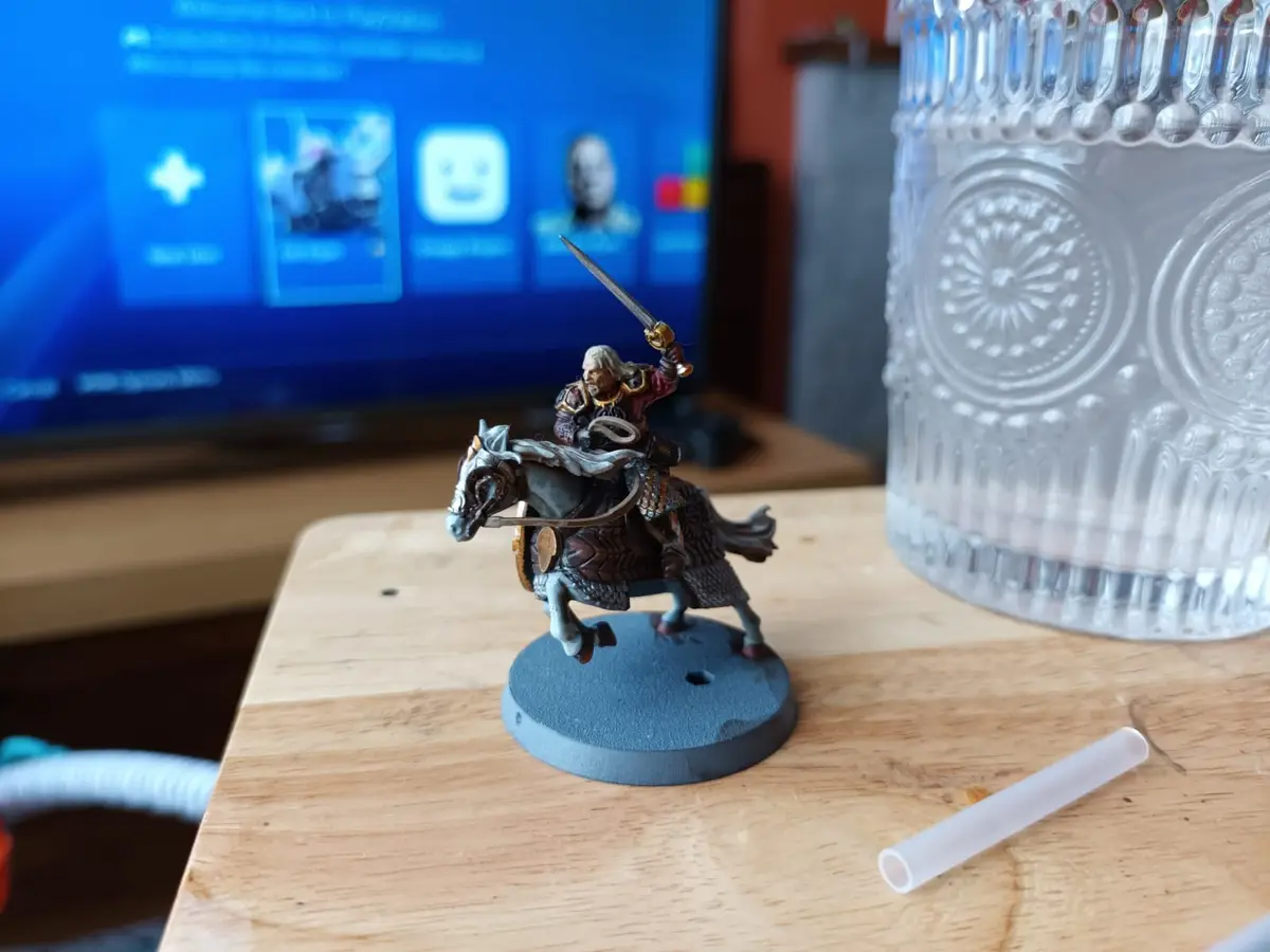 A painted miniature figure of a knight on horseback, holding a sword aloft, stands on a wooden table next to a glass and a…