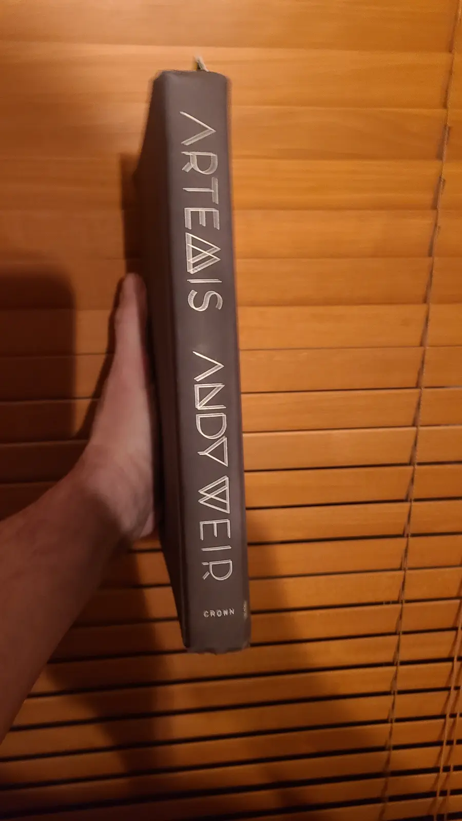 A hand holds a thick, dark gray book upright against wooden blinds, showing its spine which reads 'ARTEAIS ANDY WEIR' in…