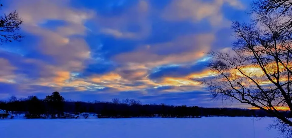 A serene winter landscape featuring a snow-covered field, silhouetted bare trees, and a dramatic sky with deep blue clouds…