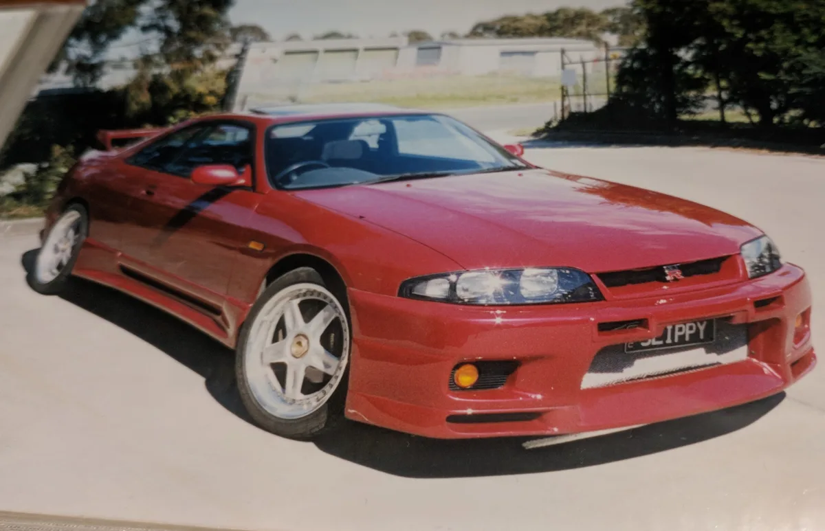 A red Nissan Skyline R34 sports car with aftermarket wheels and a black stripe is parked outdoors, its license plate reads…