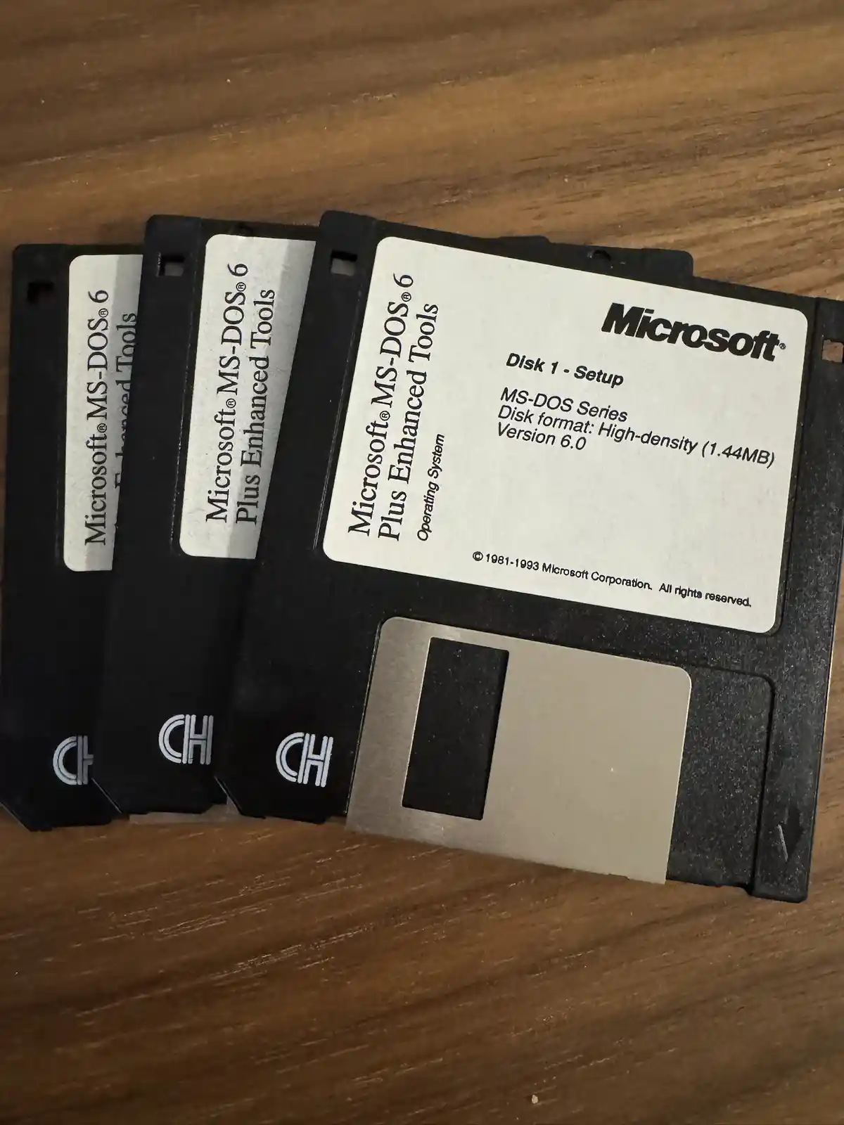 Three black 3.5-inch floppy disks labeled 'Microsoft MS-DOS 6 Plus Enhanced Tools' are fanned out on a wooden surface. The…