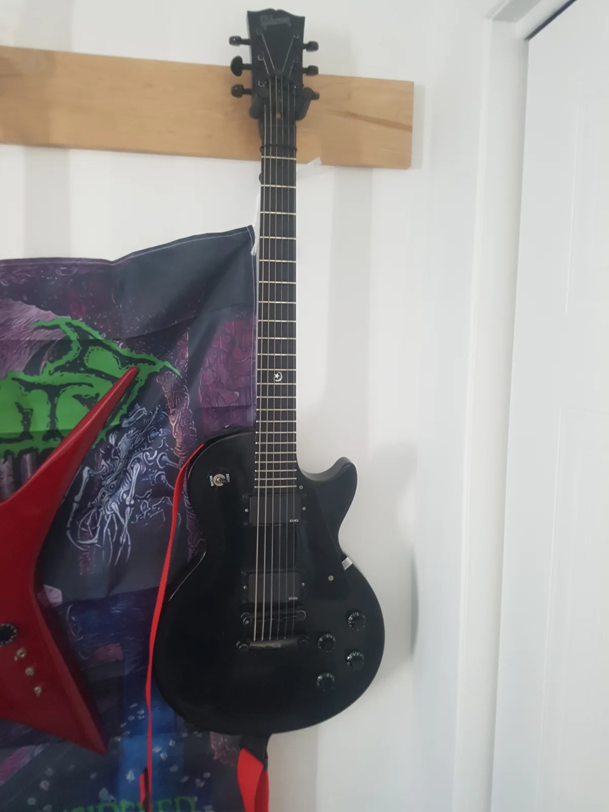 A black electric guitar hangs vertically on a wooden wall mount next to a red guitar and a dark, colorful poster with green…