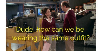 Two people in a TV studio or newsroom, both wearing identical maroon sweaters and light-colored pants, stand facing each…