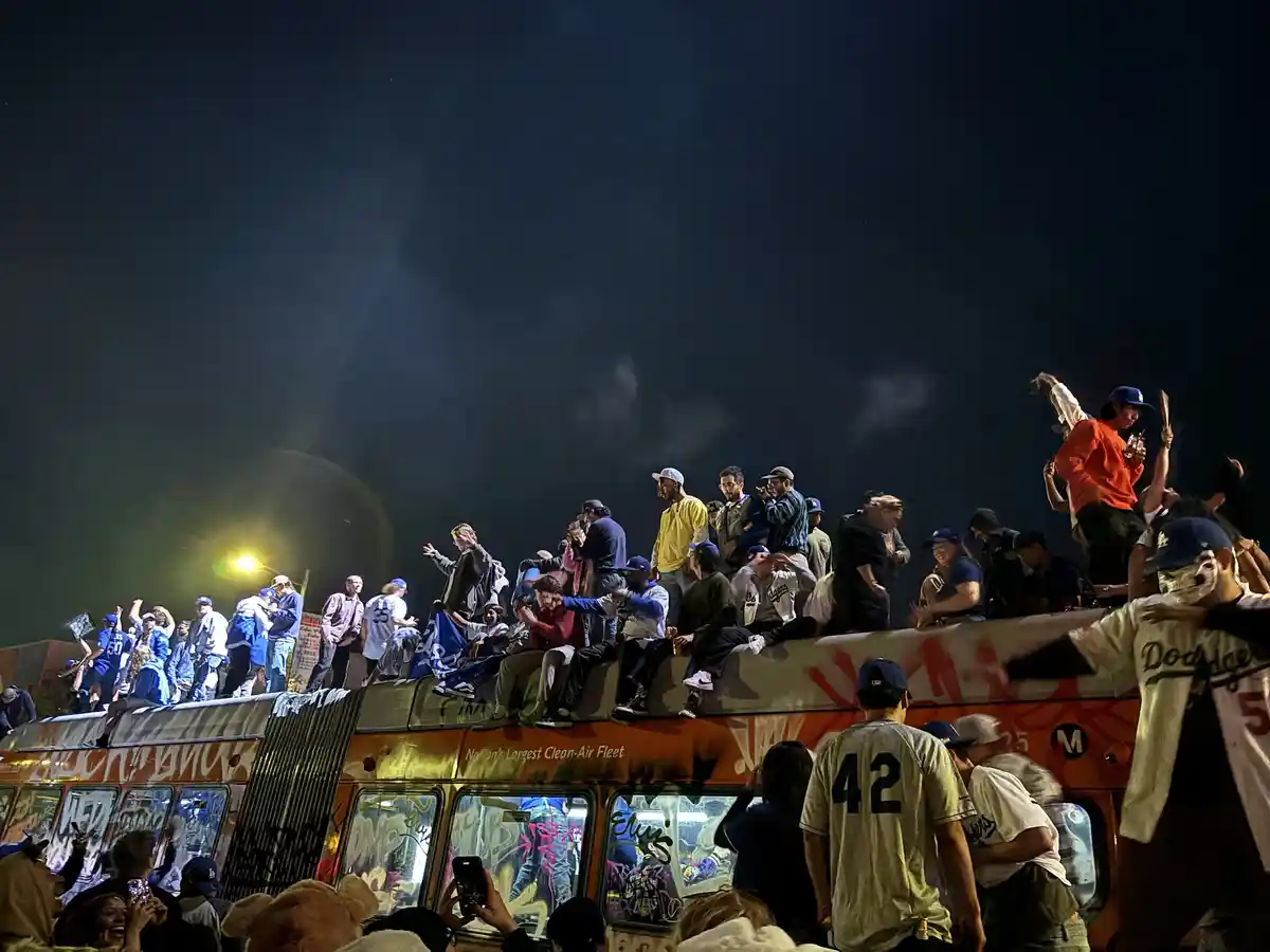 A nighttime photo shows a crowd of people, many wearing Dodgers baseball jerseys and caps, celebrating atop a…