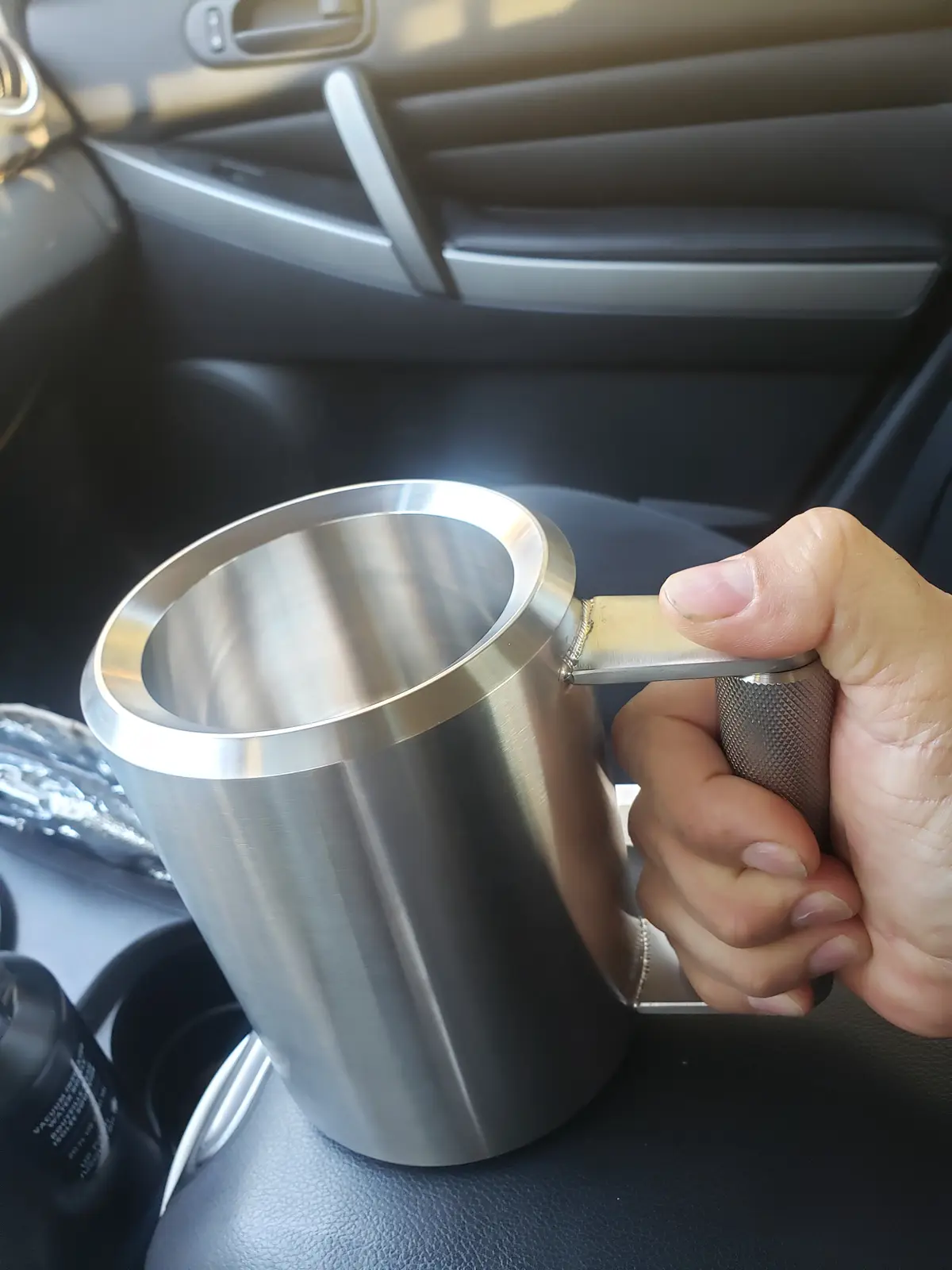A hand grips a stainless steel travel mug inside a car, with the mug's handle visibly welded or crudely attached. The…