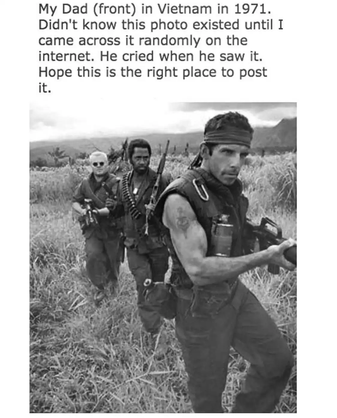 A black-and-white photo shows three men in military gear walking through tall grass in a field. The man in the foreground…