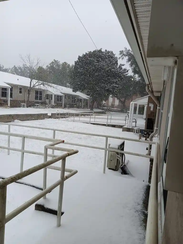 A snowy outdoor scene viewed from a porch, showing snow-covered ground, railings, and buildings under a gray sky, with the…