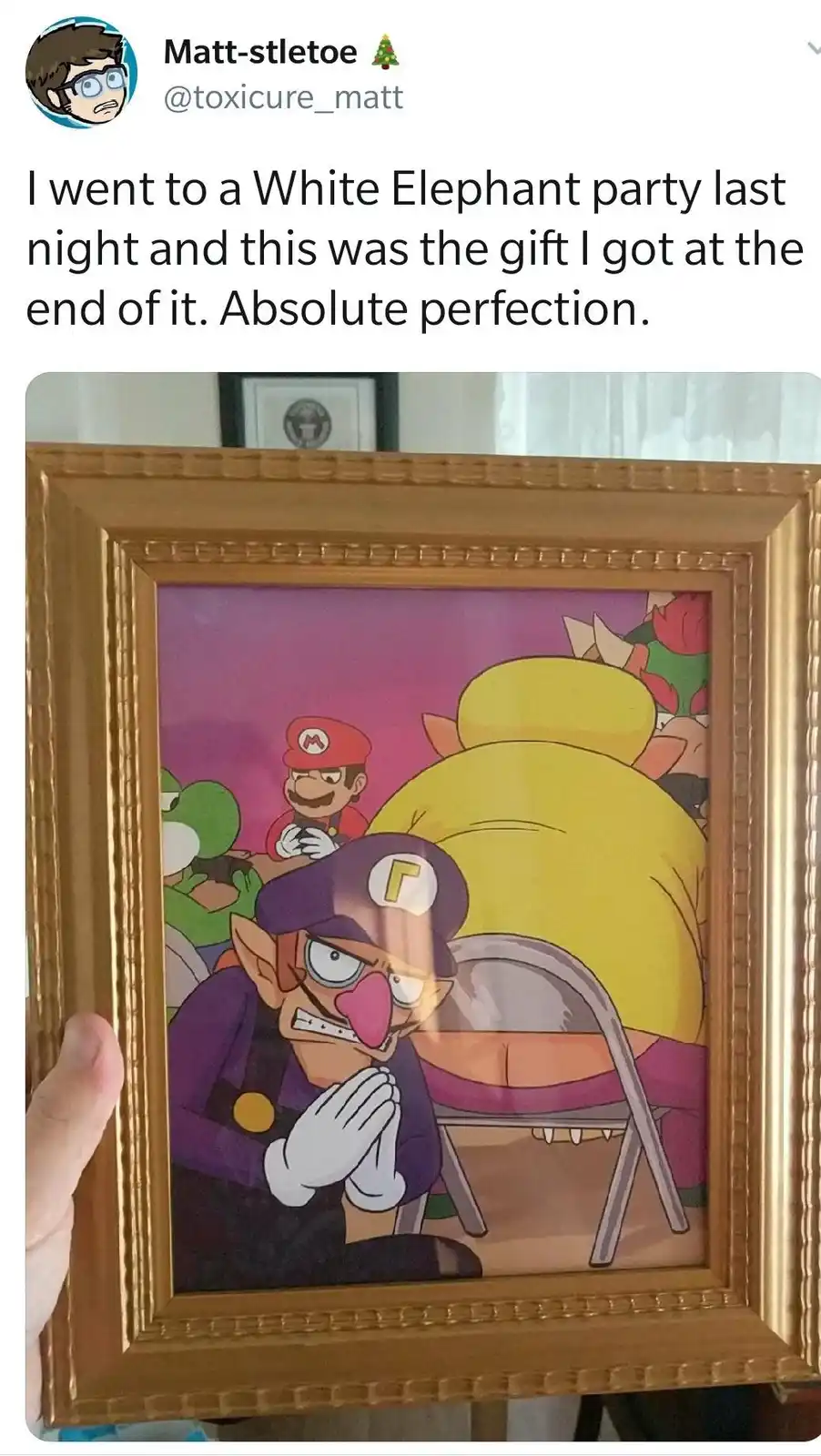 A person holds up a gold-framed picture showing a cartoon scene where a character resembling Waluigi is kneeling with…