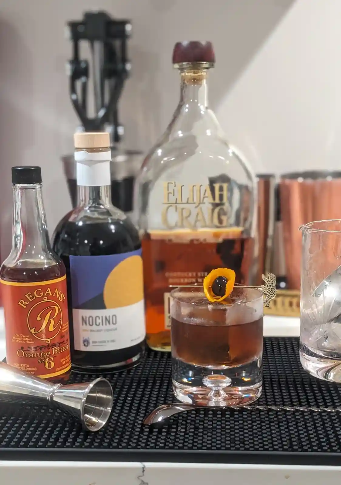 A close-up photo of a cocktail bar setup featuring a finished drink in a rocks glass garnished with an orange twist and…