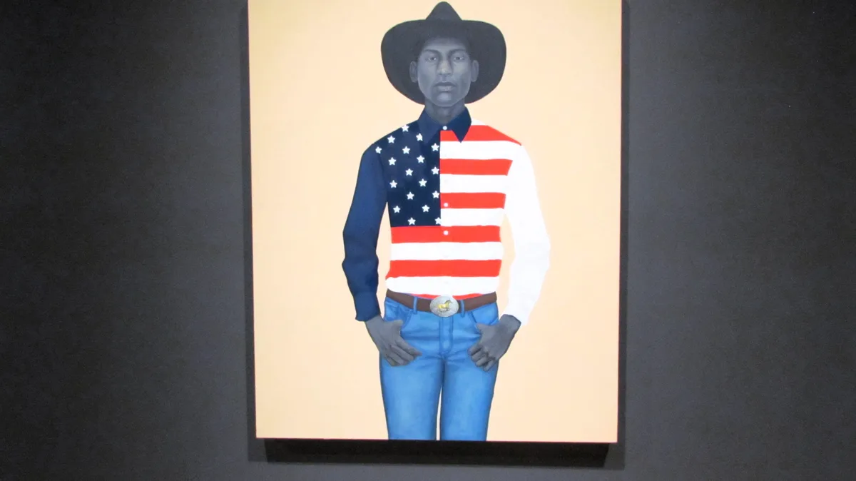 A framed painting depicts a Black man in a cowboy hat, blue jeans, and a belt with a silver buckle, standing with hands in…