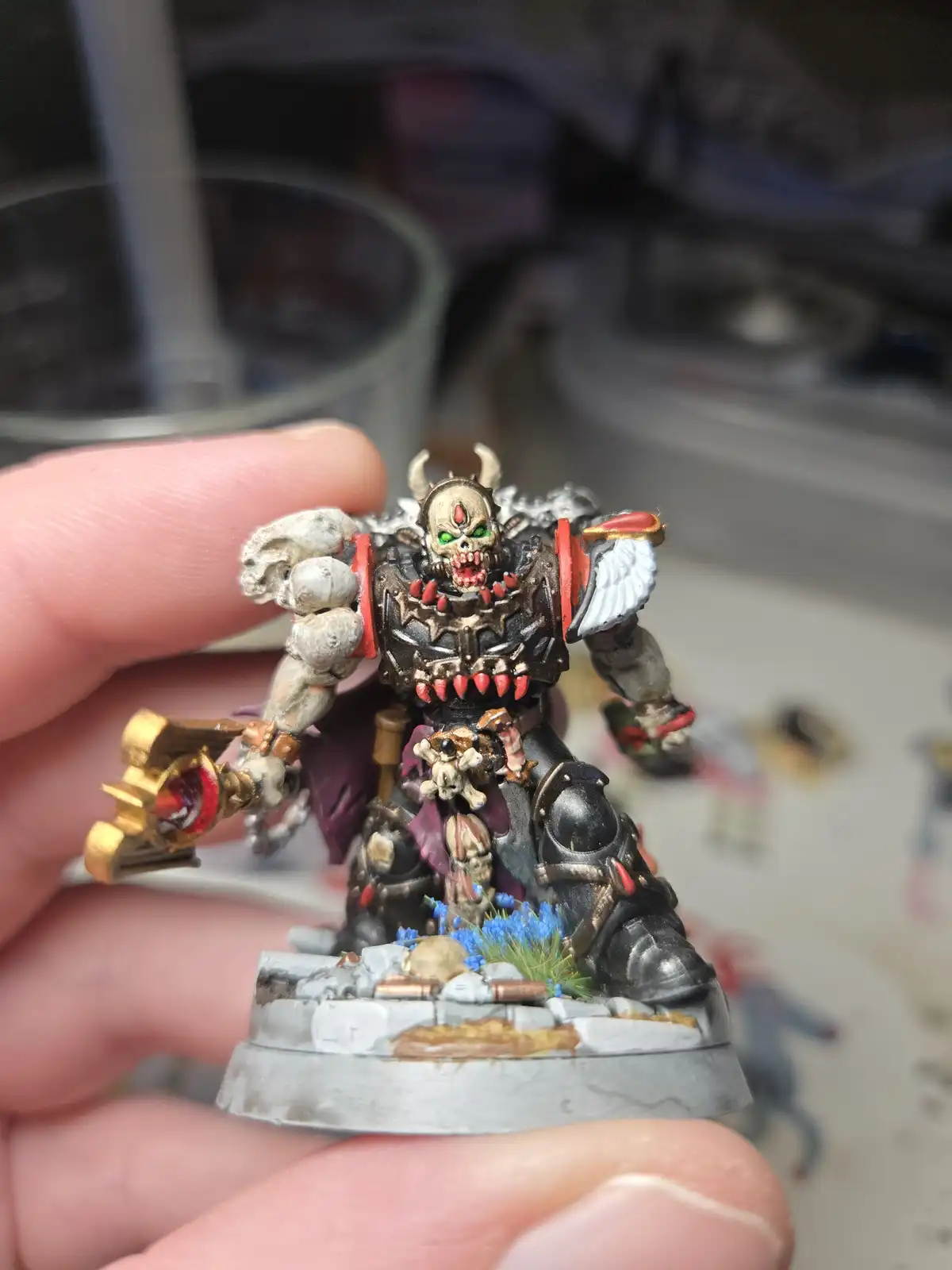 A hand holds a highly detailed, painted miniature of a grimdark fantasy warlord with a skull for a head, glowing green eyes…
