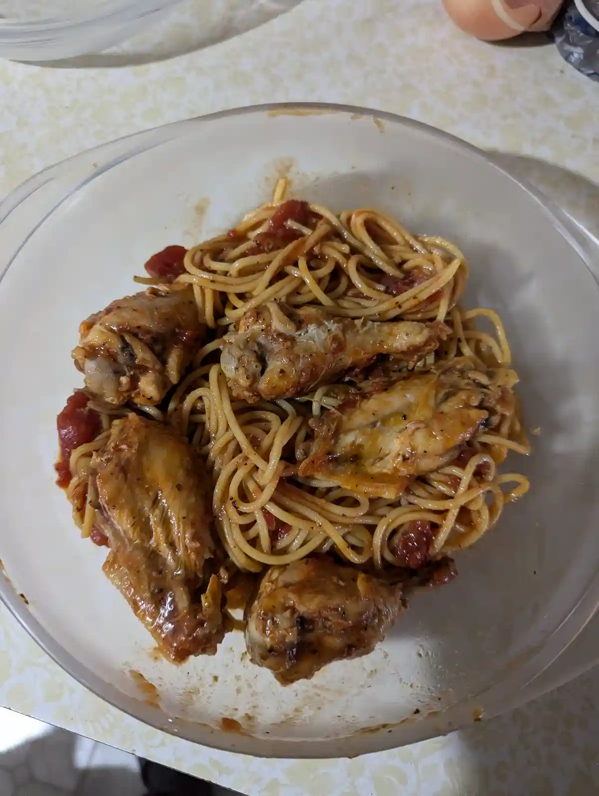 A clear glass bowl contains spaghetti with tomato sauce and several cooked chicken wings, placed on a patterned countertop…