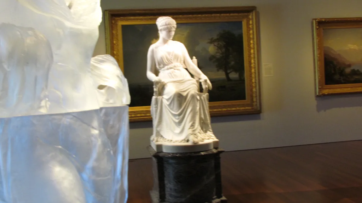 A museum photo shows a white marble statue of a seated woman in classical robes on a pedestal, with another large marble…