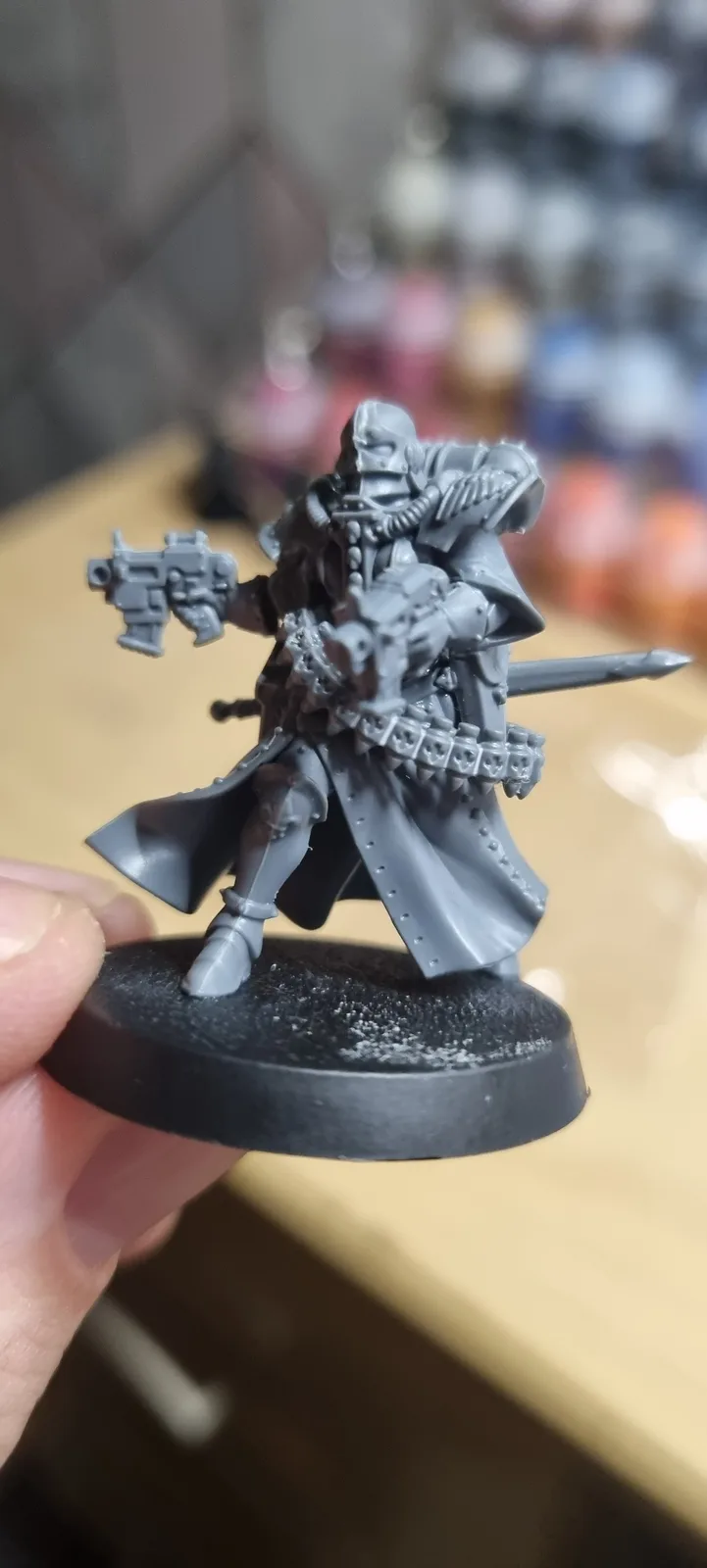 A person holds up an unpainted, grey miniature of a heavily armored warrior with a gun and sword, standing on a black base…