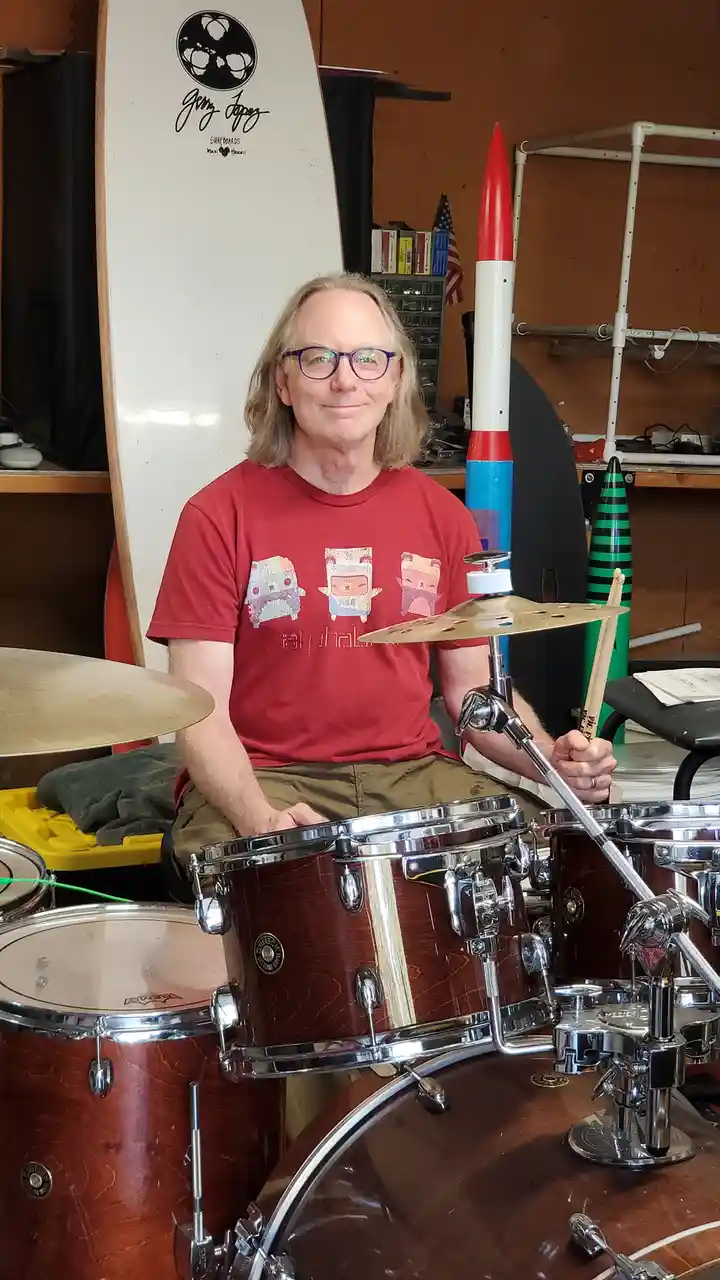 A man with glasses and a red t-shirt sits behind a drum kit, holding drumsticks. Behind him, a red, white, and blue toy…