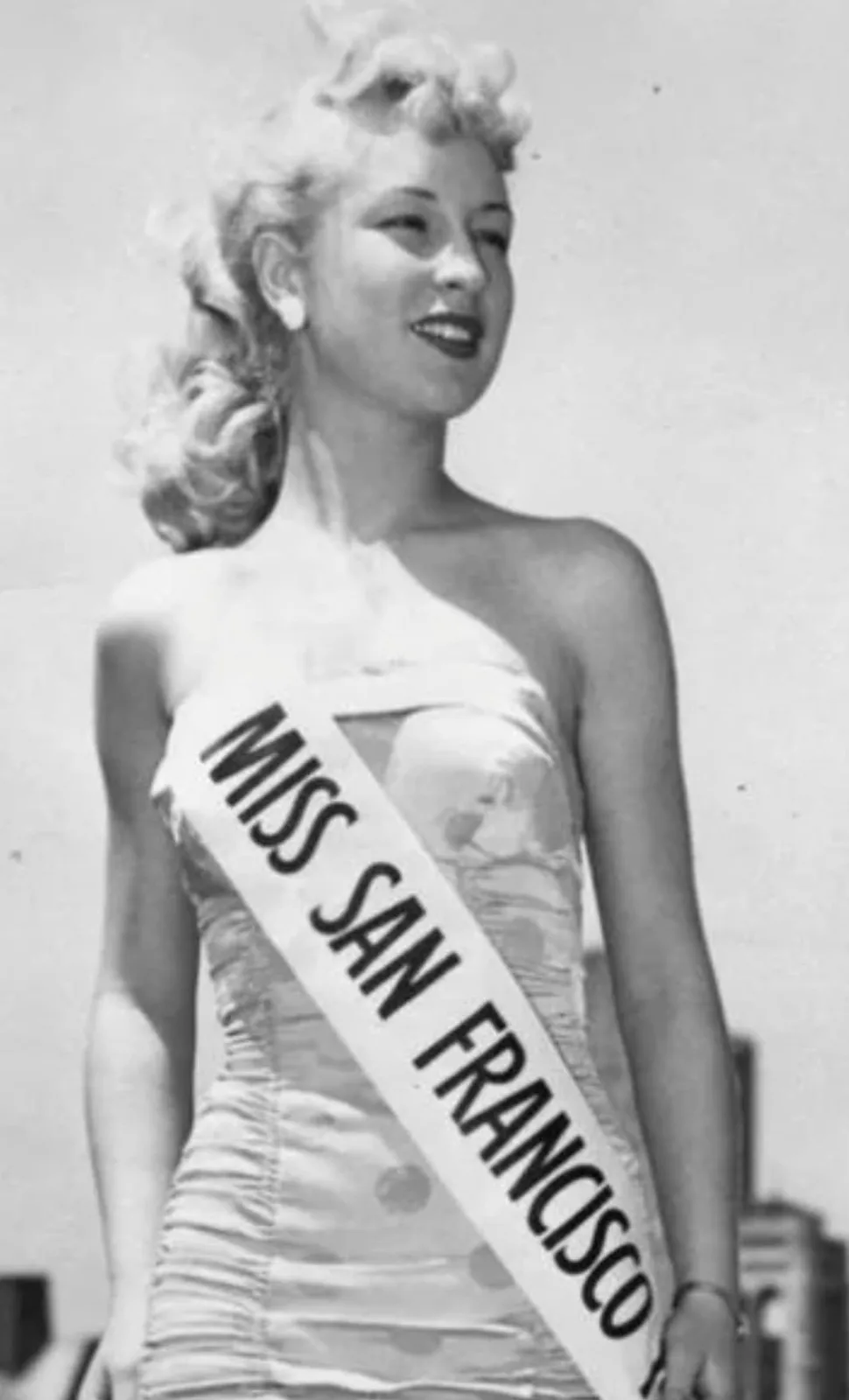 A black-and-white photograph shows a smiling woman with blonde curls wearing a strapless dress and a sash that reads 'MISS…
