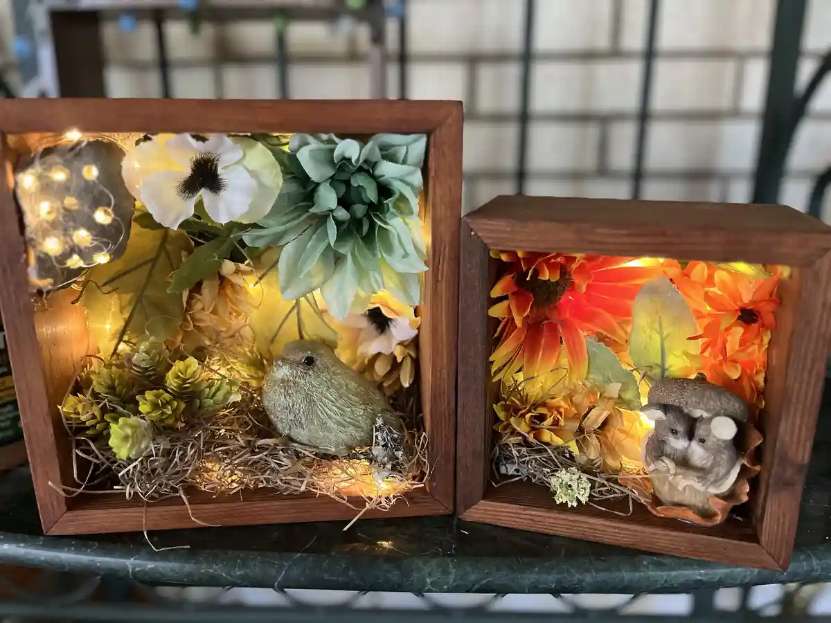 Two wooden decorative boxes with internal lighting display miniature scenes: one with a bird figurine among green and white…