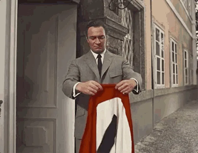 A man in a gray suit stands in a doorway holding a red, white, and black flag. He folds it carefully, then unfolds it…