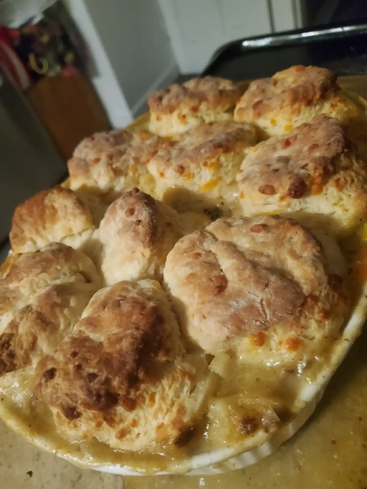 A close-up photo of a baked dish with golden-brown biscuits covering a creamy macaroni and cheese filling in a white ceramic…