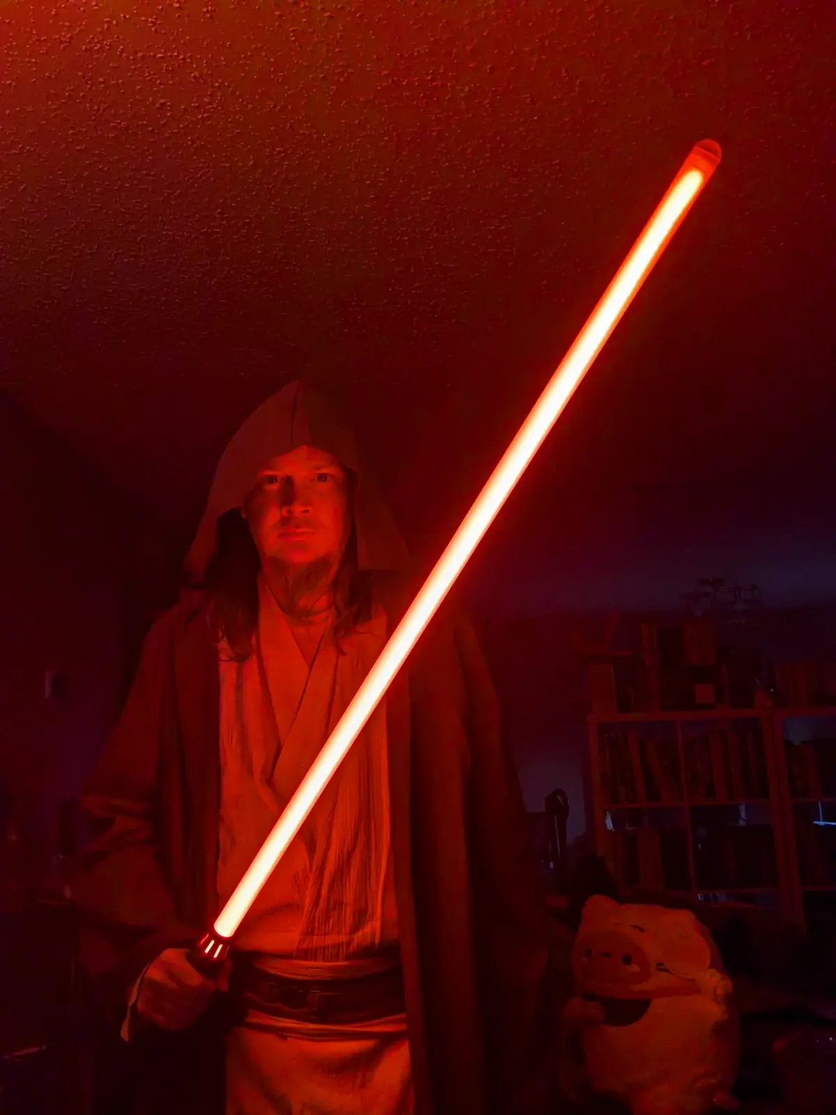 A person in a Jedi robe and hood holds a glowing red lightsaber in a dimly lit room with a textured ceiling, casting an…