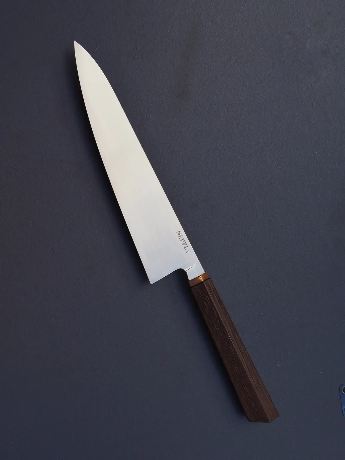 A sleek, high-quality chef's knife with a polished stainless steel blade and dark wooden handle rests diagonally on a dark…