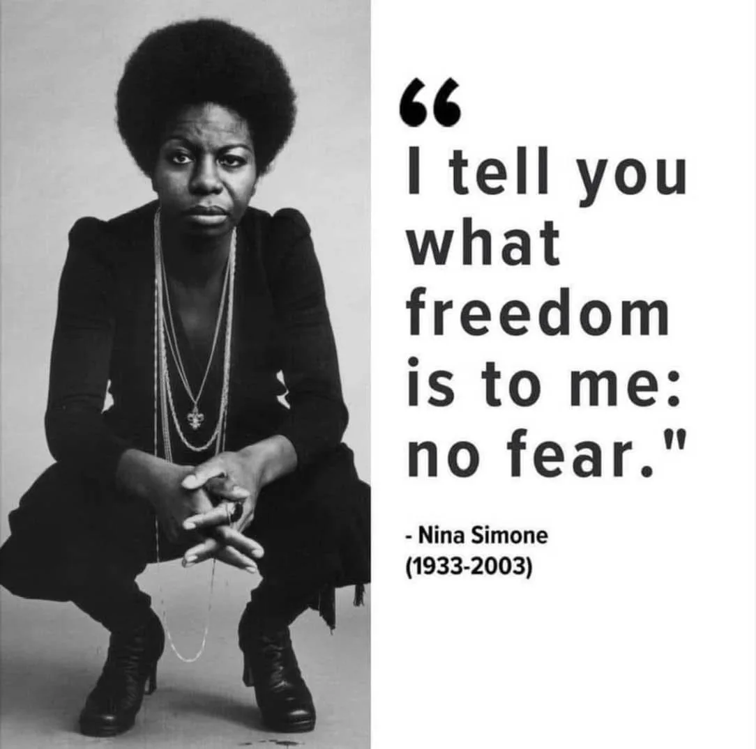 A black-and-white photo of Nina Simone crouching, looking directly at the camera with a serious expression, wearing layered…