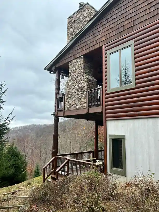 A rustic log cabin with stone chimney and wooden decks overlooks a forested hillside under an overcast sky. The overlaid…