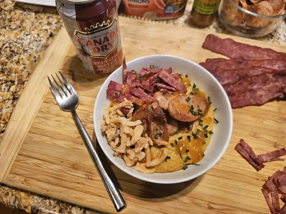 A white bowl filled with yellow grits topped with sliced sausage, crispy fried onions, and chopped green herbs, surrounded…