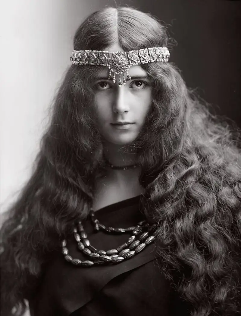 A vintage black-and-white portrait of a woman with long, wavy hair, wearing an ornate jeweled headband and layered…